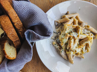 Meal in the restaurant. fresh and delicious penne pasta sprinkled with parmesan and spices. next to it is a napkin and a basket of fragrant freshly baked bread. Food is on the wooden table