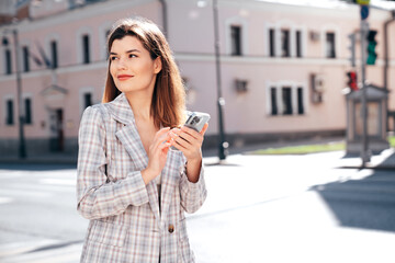 Young beautiful woman in nice trendy white jacket suit. Sexy smiling model posing in street. Fashionable female outdoors. Cheerful and happy. Holds smartphone, uses phone apps, looks at screen