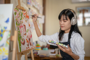A smiling young woman enjoys listens to music while painting a colorful canvas in a cozy, homey art studio.