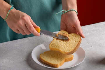 Authentic female hands spreading peanut butter on piece of white bread toast, at gray kitchen table, close-up. Typical snack food, food lifestyle, domestic life, preparing breakfast