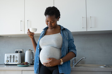 Pregnant woman holding tea cup and touching belly in kitchen