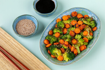 Asian-style stir-fried broccoli with carrot. Dark sauce and roasted sesame seeds in small bowls. Bamboo mat and chopsticks.