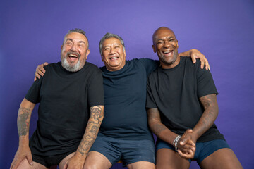Portrait of three men laughing while sitting on bench against purple background