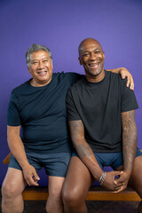 Portrait of two men laughing while sitting on bench against purple background