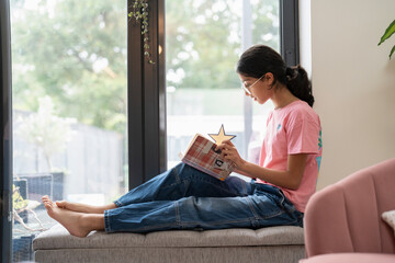 Girl sitting at hassock near window and reading book