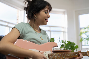 Young woman sitting on sofa and playing guitar - obrazy, fototapety, plakaty