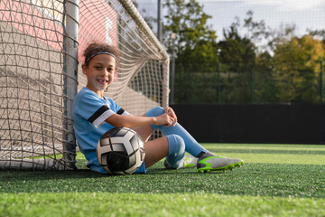 Portrait of girl (6-7) with ball sitting next to soccer goal