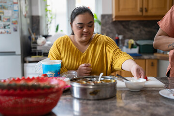 Young woman with down syndrome preparing food in kitchen - obrazy, fototapety, plakaty