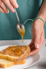 Woman's hands puting honey with a spoon on a toast with peanut butter to make a sandwich for breakfast, at gray kitchen table, close-up. Honey dripping from a spoon onto a peanut butter sandwich