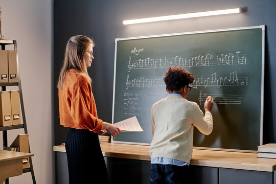 Young Caucasian teacher of music watching her Black student writing notes on staff on blackboard during lesson