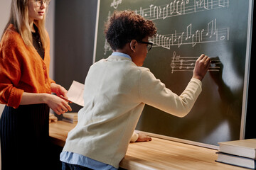 African American boy writing notes blackboard during music class, his Caucasian female teacher...