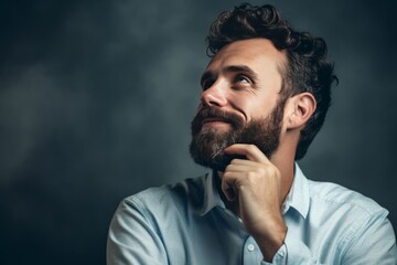 Smiling bearded man looking up with hand on chin on dark moody background. The thoughtful look is lost in the distance. Copy space.