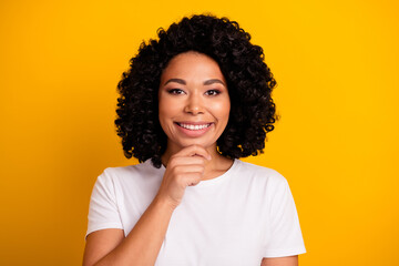 Portrait of toothy beaming positive girl with curly hairdo wear white t-shirt smiling hand on chin isolated on yellow color background