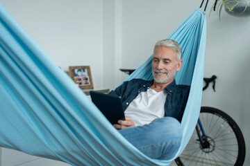 Smiling mature man using digital tablet in hammock - obrazy, fototapety, plakaty