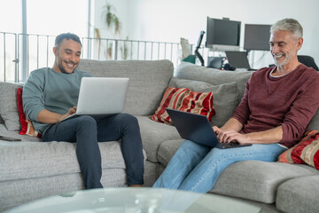 Smiling male couple working on laptops on sofa at home