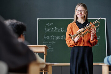Medium long shot of cheerful young music teacher holding trumpet standing in classroom working with...