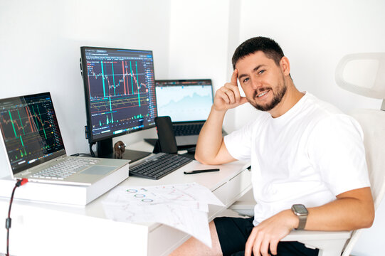 Photo of a confident successful male trader sitting at desk at home office monitoring stock market, analyzing price flow, risks and prospects, smiles at camera. Stock trading, cryptocurrency concept
