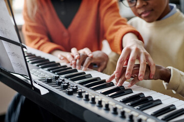 Selective focus shot of unrecognizable woman teaching Black boy to play synthesizer at music class
