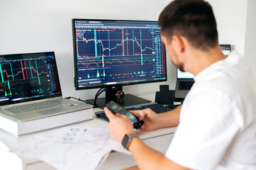 Fototapeta na wymiar Male trader sits in home office in front of screens with graphs and financial charts, using smartphone, predicts, analyzing and studying the growth or decline of cryptocurrency.Stock exchange, trading