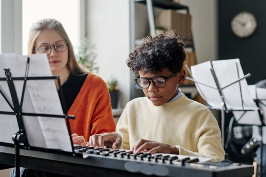 African American boy practicing synthesizer at music class a school, young Caucasian teacher sitting next to him
