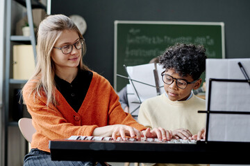 Young Caucasian woman wearing eyeglasses teaching African American boy to play synthesizer at music...