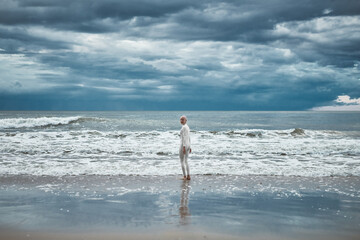 Hairless girl with alopecia in white futuristic suit goes to cold restless sea on sandy beach,...