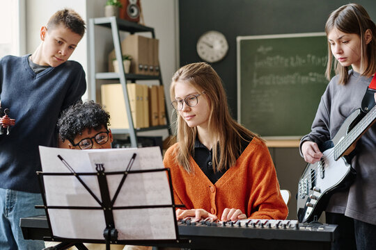 Young female teacher playing synthesizer during music class at school, her students sitting and standing next to her