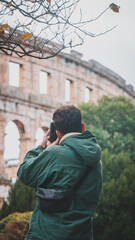 White man taking a picture with his camera to the roman Arena in Pula, region of Istria in Croatia, and wearing a green jacket on a cloudy day