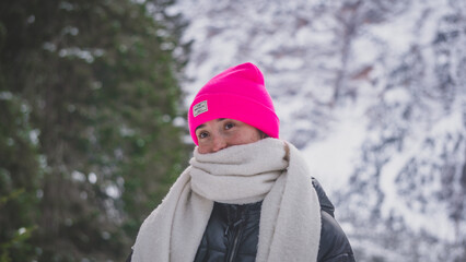 Young white woman wearing a pink winter hat and a white scarf looking to the someone else's camera while travelling to Lago di Braies in the Dolomites, Itlay, during a winter and snowy day