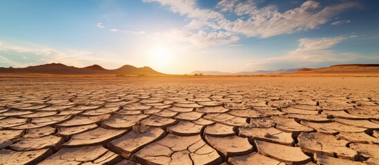 The sun is setting over a barren desert, casting a warm orange glow over the dry, cracked landscape. The horizon is painted with hues of red and purple as the day transitions into night, highlighting