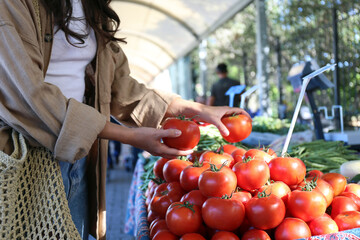 Young woman with reusable eco friendly net bag picking fresh tomatoes on farmers market. Conscious...