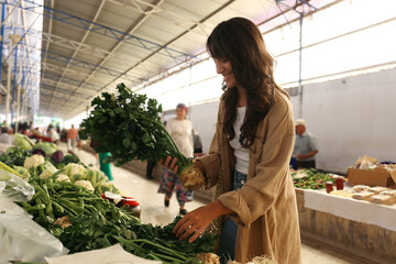 Young woman with reusable eco friendly net bag picking out fresh celery roots on farmers market....