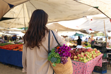 Middle eastern young woman at farmers market with an eco friendly straw bag full of healthy produce. Conscious shopping for organic fruits and vegetables. Close up, copy space, background.