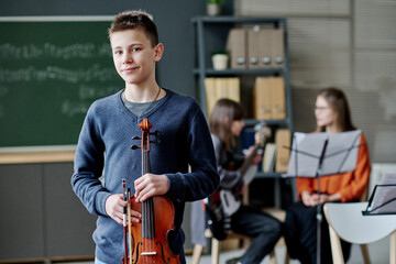 Medium portrait of confident teen boy holding violin standing in classroom looking at camera, his...