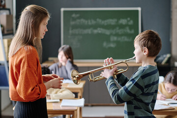 Schoolboy standing in front of young music teacher practicing trumpet during class at school,...