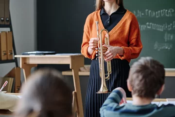 Poster Selective focus crop shot of unrecognizable female music teacher holding brass instrument standing in front of kids in classroom © AnnaStills