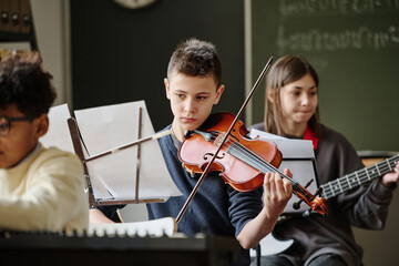 Selective focus of boy reading sheet music and playing violin during practice with school orchestra