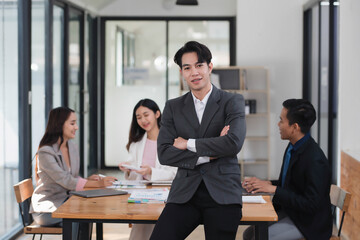 Fototapeta na wymiar Young confident Asian businessman standing in the office room with their coworkers in the background.