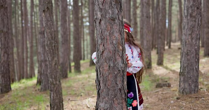 Bulgarian woman young girl in ethnic folklore embroidery costume hiding behind tree trunk in the forest in mountains, Bulgaria