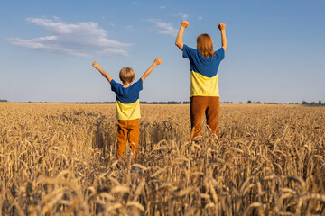 woman and a child stand with their backs in identical yellow and blue T-shirts among a wheat field...