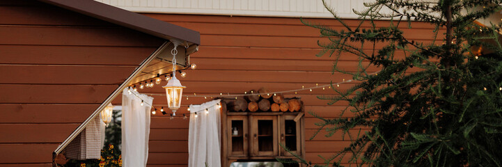 Cozy, illuminated wooden home exterior with a white picket fence. The brown wooden house is adorned with hanging lights, welcoming ambiance. Banner with copy space.