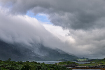 A dynamic summer sky in Norway stretches over a rugged landscape, where mountains and foliage are caressed by rolling mist, near the Arctic Circle