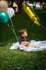 Bebé de un año sentado sobre una manta blanca en un parque iluminado con la luz del día junto a varios globos de colores y un globo dorado del número uno