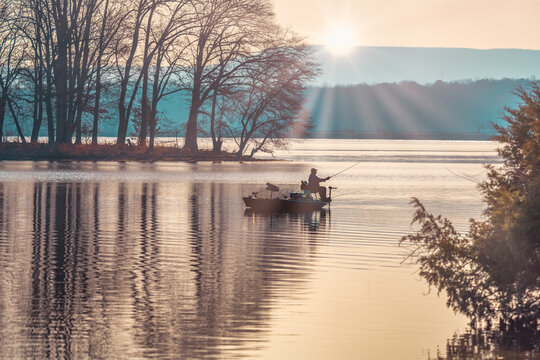 Man crappie fishing in a bass boat on Woods Reservoir Lake in Tennessee with early morning sunrise over the mountain silhouette.