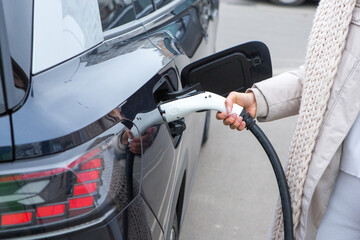 Young woman charging her electric car at the gas station, using a smartphone. Eco fuel concept. The...