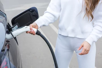 Fotobehang Young woman charging her electric car at the gas station, using a smartphone. Eco fuel concept. The concept of environmentally friendly transport. Recharging battery from charging station. © romeof