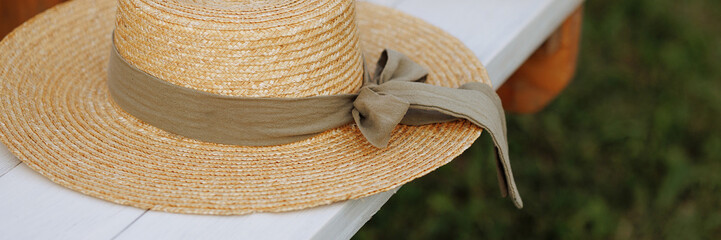 A straw hat with a ribbon rests on a white bench amidst greenery. Concept: summer fashion, outdoor relaxation, garden aesthetics. Banner with copy space.