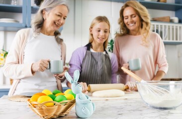 Three generations unite in kitchen, mother, grandmother, and granddaughter, as they preparing for Easter with love and laughter. Basket with painted eggs and Easter bunny in foreground