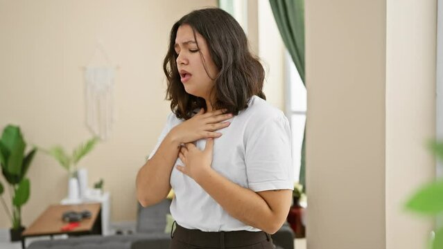 Unwell young beautiful hispanic woman coughing at home, standing in the living room, signaling health care for suspected cold or bronchitis symptoms.