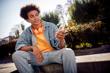 Obraz premium Photo of good mood youngster with afro hairdo dressed jeans jacket headphones sitting on platform texting email on smartphone outdoors